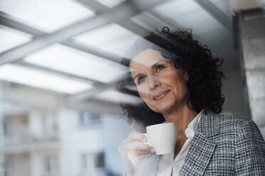 Mature female professional with coffee cup contemplating while looking through window - MOEF03869