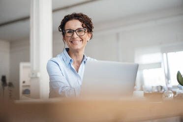 Lächelnde Geschäftsfrau mit Brille sitzt mit Laptop im Büro - MOEF03877