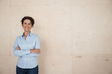 Smiling mature female professional standing with arms crossed in front of beige wall - MOEF03881