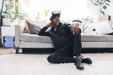 Entspannter Mann mit Virtual-Reality-Headset, der wegschaut, während er sich zu Hause auf dem Sofa ausruht - ASGF01083