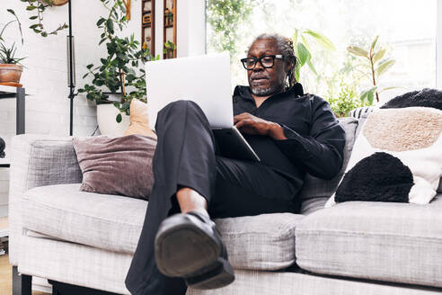 Älterer männlicher Berufstätiger, der einen Laptop benutzt, während er zu Hause auf dem Sofa sitzt - ASGF01097