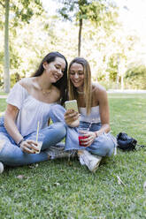 Junge Freundinnen teilen sich ein Smartphone, während sie in einem öffentlichen Park sitzen - JRVF01434