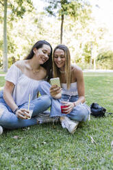 Young female friends sharing smart phone while sitting at public park - JRVF01434