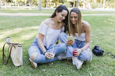 Freundinnen halten Getränke in der Hand und teilen sich ein Smartphone in einem öffentlichen Park - JRVF01435