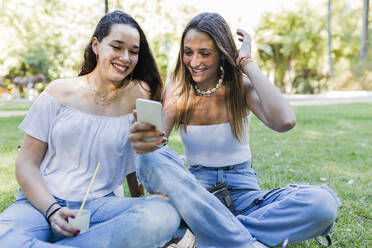 Lächelnde Frau, die mit ihrer Freundin im Park sitzt und ein Smartphone benutzt - JRVF01438