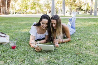 Junge Freundinnen teilen sich ein digitales Tablet, während sie im Park im Gras liegen - JRVF01441