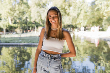 Smiling beautiful blond woman standing in front of pond at public park - JRVF01446