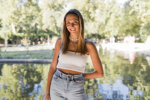 Lächelnde schöne blonde Frau steht vor einem Teich in einem öffentlichen Park - JRVF01446