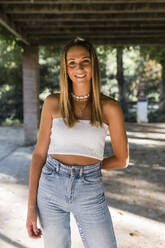 Smiling young blond woman standing at public park - JRVF01448
