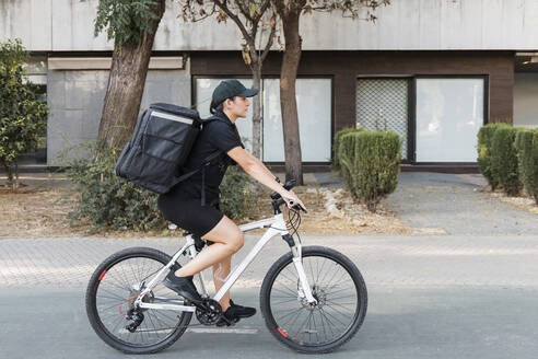 Weibliche Lieferperson mit Rucksack auf dem Fahrrad auf der Straße - JRVF01460