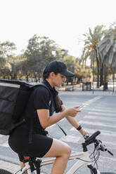 Junge Zustellerin, die auf dem Fahrrad sitzend ein Mobiltelefon benutzt - JRVF01466