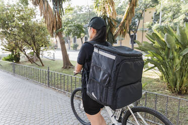 Weiblicher Zusteller, der einen Rucksack trägt, während er mit dem Fahrrad auf dem Gehweg fährt - JRVF01469