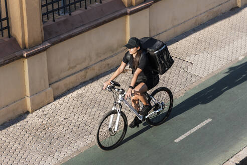 Weibliche Lieferperson fährt Fahrrad auf der Straße an einem sonnigen Tag - JRVF01481