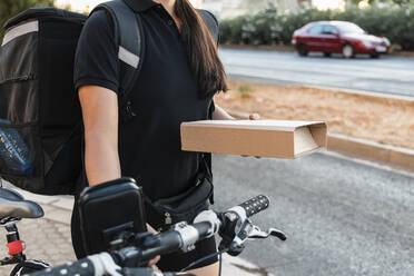 Weiblicher Angestellter, der ein Kurierpaket hält, während er mit dem Fahrrad fährt - JRVF01496