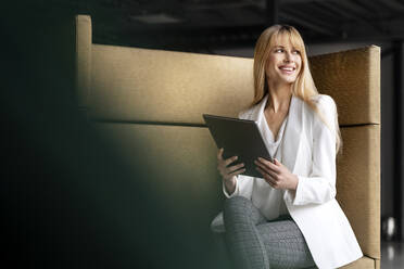 Lächelnde Geschäftsfrau, die wegschaut, während sie mit einem digitalen Tablet auf dem Sitz sitzt - PESF03083