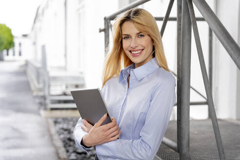 Lächelnde Geschäftsfrau, die ein digitales Tablet hält und sich auf ein Metallgeländer stützt - PESF03110