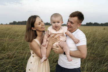 Fröhliche Mutter und Vater, die gemeinsam ihre Tochter auf einem Feld tragen - EYAF01711