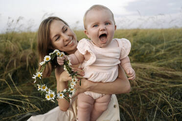 Lächelnde Mutter, die ein fröhliches kleines Mädchen mit einem Blumen-Diadem im Feld hält - EYAF01713