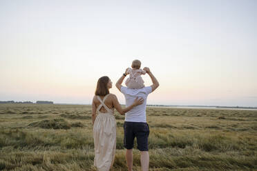 Father carrying baby girl on shoulders while walking with woman in field - EYAF01716