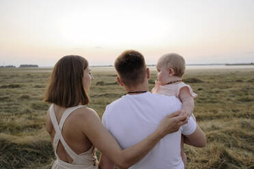 Frau hält die Hand ihrer Tochter, während sie neben einem Mann auf einem Feld bei Sonnenuntergang steht - EYAF01718