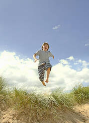 Carefree boy jumping over sand dunes at beach during sunny day - AJOF01583