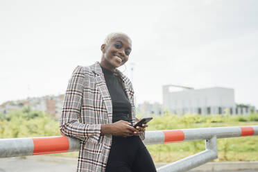 Smiling woman holding mobile phone while leaning on railing - MEUF03954