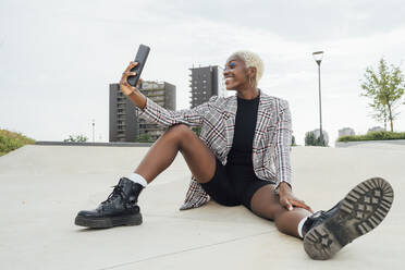 Smiling woman taking selfie through smart phone while sitting on footpath - MEUF03959