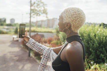 Junge Frau nimmt Selfie durch Handy im Park an einem sonnigen Tag - MEUF03967