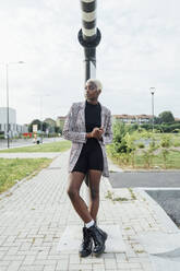 Woman with hands clasped leaning on pole while standing on footpath - MEUF03973