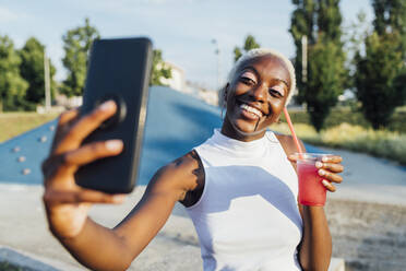 Smiling woman taking selfie through smart phone while holding smoothie at park - MEUF03998