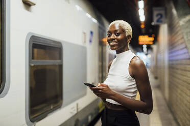 Smiling woman holding mobile phone while standing at subway station - MEUF04015