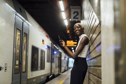 Lächelnde Frau mit Mobiltelefon an der Wand lehnend in einer U-Bahn-Station - MEUF04017