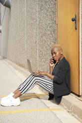 Businesswoman talking on smart phone while sitting with laptop on footpath - VEGF04858