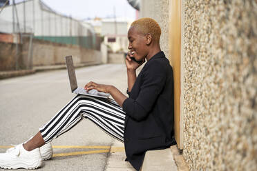 Junge Geschäftsfrau, die am Laptop arbeitet, während sie auf dem Gehweg mit ihrem Handy telefoniert - VEGF04860