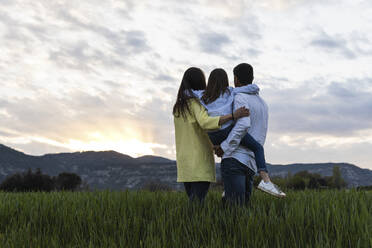 Vater und Mutter, die ihre Tochter tragen, während sie bei Sonnenuntergang die Aussicht von einem Feld aus betrachten - PNAF02114