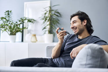 Lächelnder junger Mann, der eine Sprachnachricht auf seinem Mobiltelefon sendet, während er zu Hause auf dem Sofa sitzt - BSZF01815