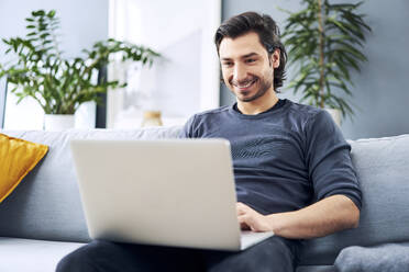 Businessman working on laptop while sitting on sofa at home - BSZF01820