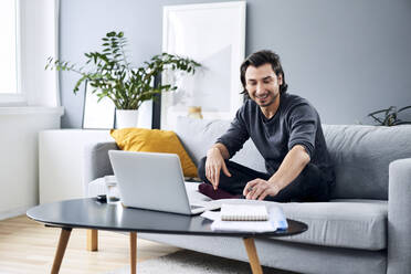 Lächelnder männlicher Berufstätiger bei der Arbeit, während er zu Hause auf dem Sofa sitzt - BSZF01836