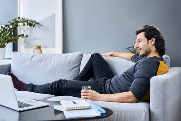 Businessman relaxing on sofa during break while working at home - BSZF01839