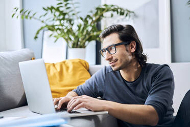 Männlicher Student mit Brille, der zu Hause einen Laptop benutzt - BSZF01843