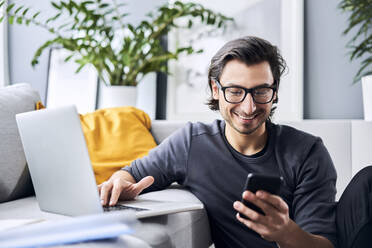 Lächelnder Mann, der ein Smartphone benutzt, während er zu Hause am Laptop tippt - BSZF01845