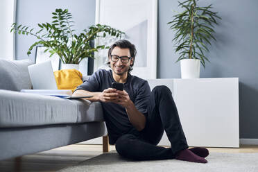 Lächelnder Geschäftsmann, der sein Smartphone benutzt, während er sich zu Hause auf dem Sofa ausruht - BSZF01847