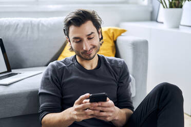 Man using smart phone while leaning on sofa at home - BSZF01849