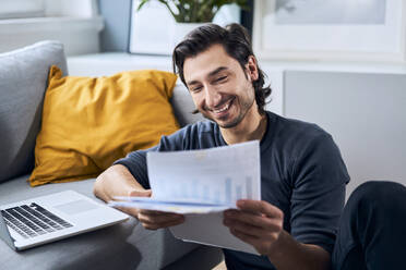 Smiling businessman looking at documents while leaning on sofa at home - BSZF01857