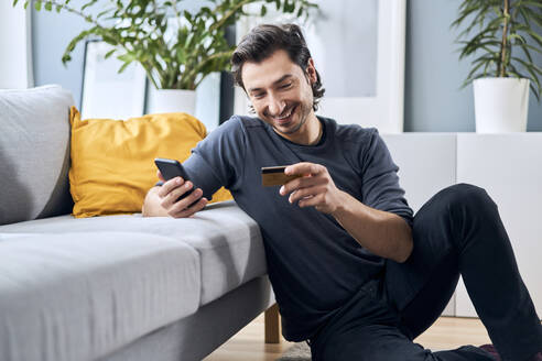 Smiling man with smart phone shopping online through credit card at home - BSZF01863