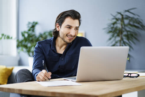 Businessman holding pen while working on laptop at home office - BSZF01882