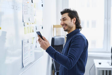 Männlicher Webdesigner benutzt ein Smartphone in der Nähe eines Whiteboards im Büro - BSZF01897