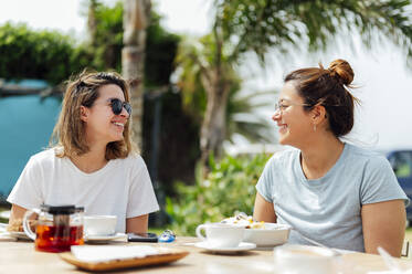 Smiling female friends looking at each other while having breakfast - PGF00741