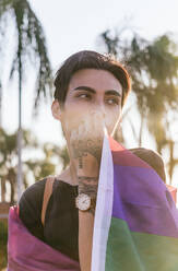 Tranquil gay male with tattoos wrapped in colorful LGBT flag in city street looking away - ADSF28414