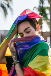 Side view of tranquil gay male with closed eyes wrapped in colorful LGBT flag in city street - ADSF28416